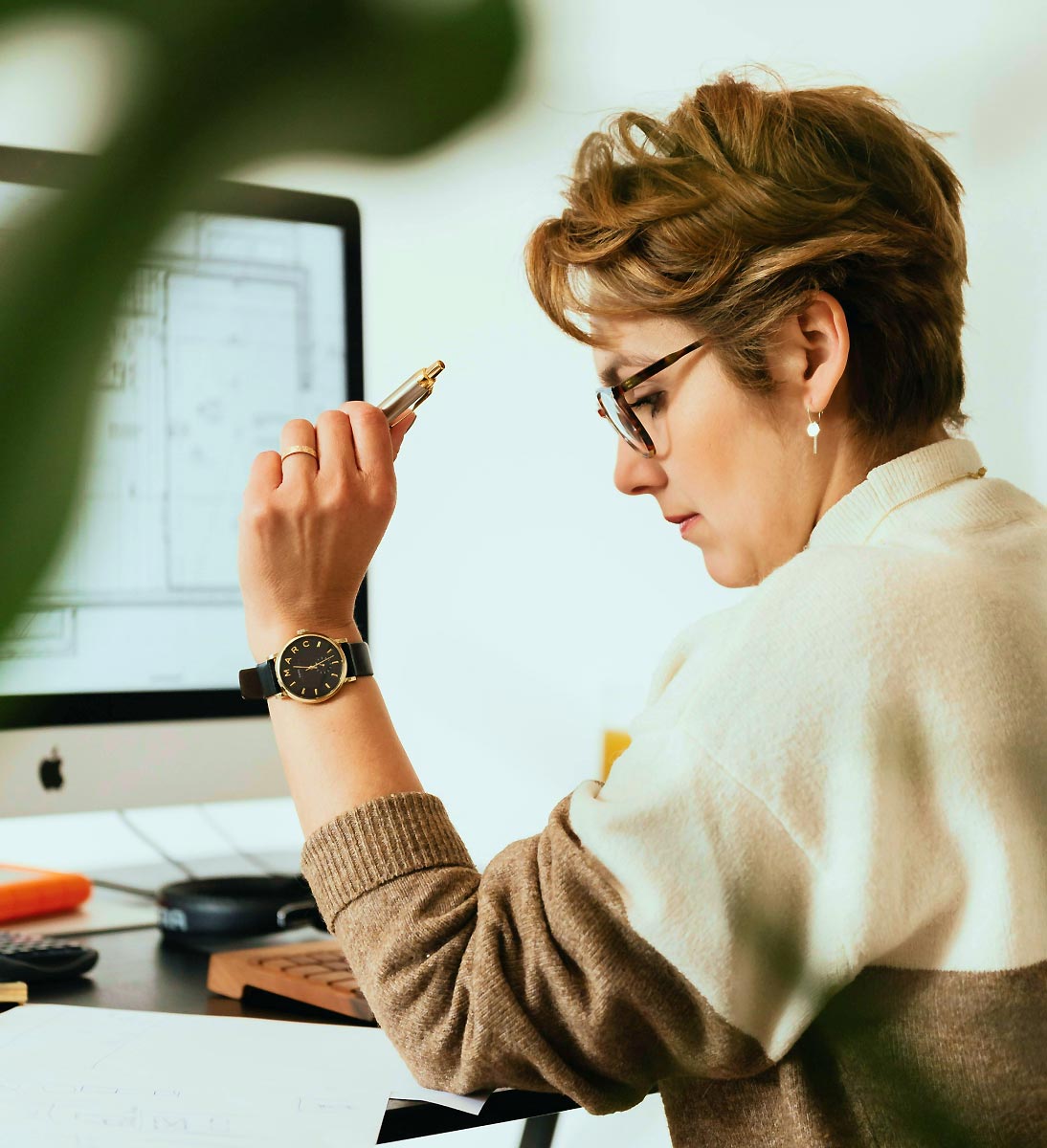 Frau am Schreibtisch Eine Frau mit kurzem braunen Haar sitzt vor einem IMac am Schreibtisch, grübelt und hält einen Kugelschreiber in der Hand.