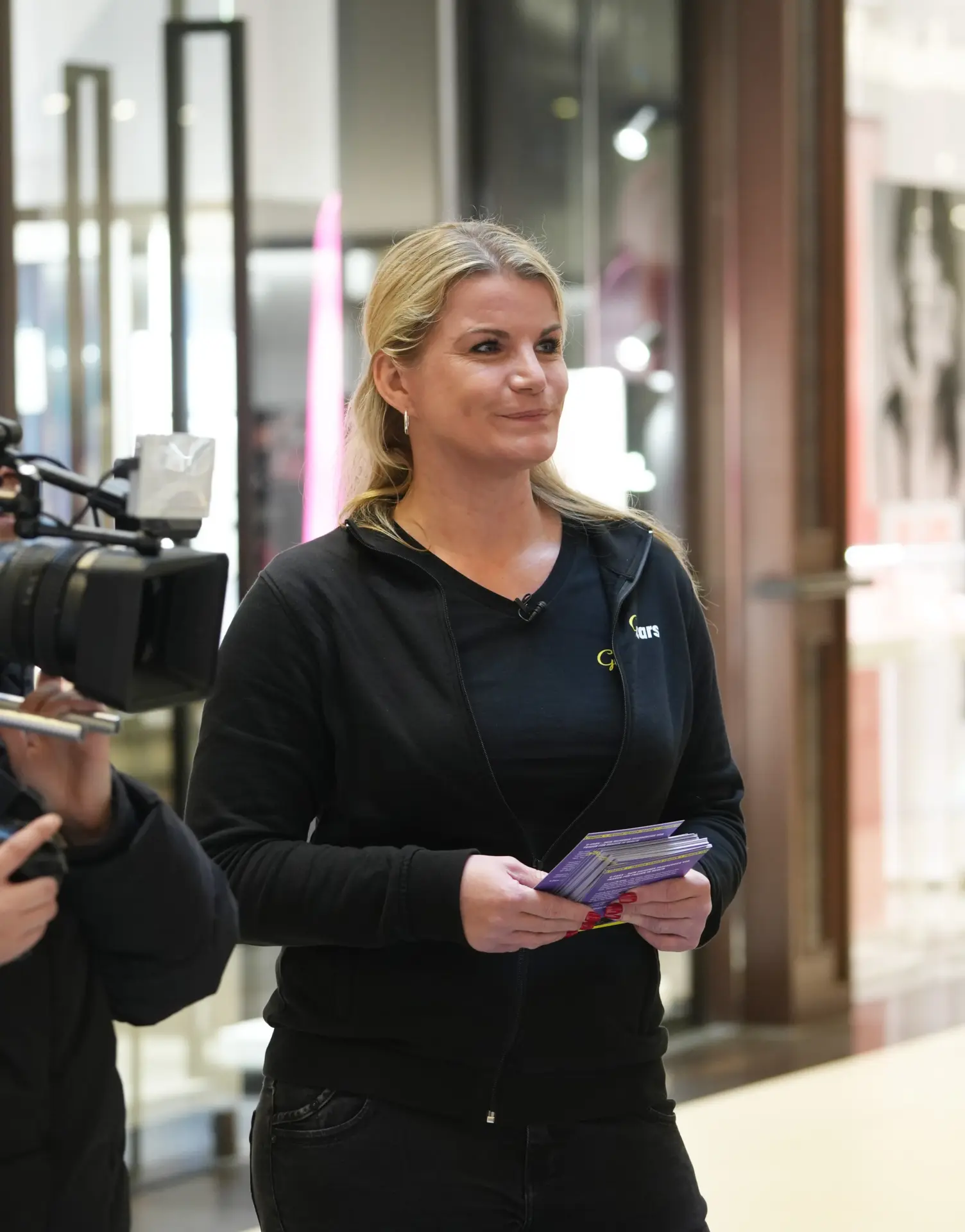 Nadin Guener steht neben der Video-Kamera, hält Bilder in der Hand, lächelt und schaut nach links.
